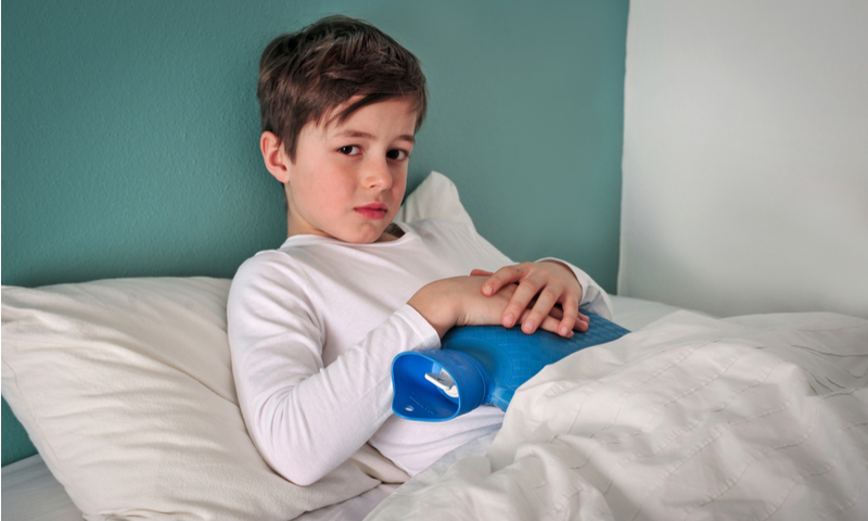 Niño pequeño enfermo en la cama sujetando una bolsa de agua caliente contra su abdomen