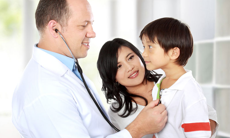 Médico auscultando el corazón de un niño pequeño mientras su madre está de pie junto a ellos.