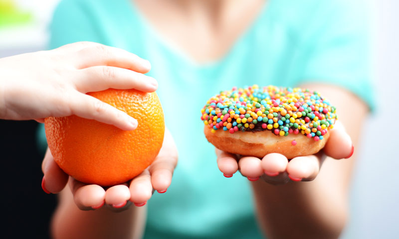 Madre sosteniendo una naranja en una mano y una rosquilla en la otra, la mano del niño se estira para tomar la naranja