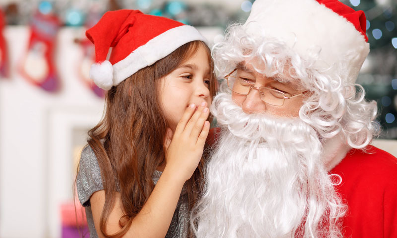 Niña pequeña en el regazo de Santa Claus susurrándole lo que quiere en el oído