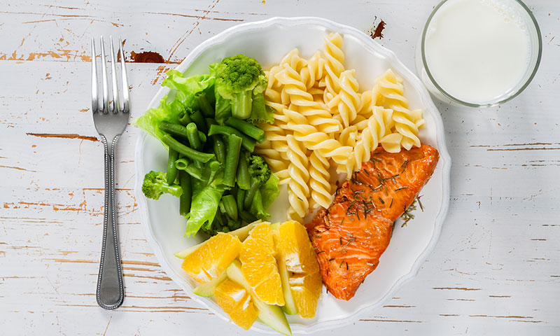 Plato con porciones saludables de salmón, pasta, verduras, frutas y leche
