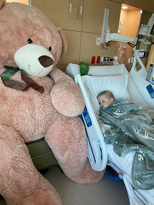 Young boy laying in hospital bed with large teddy bear.