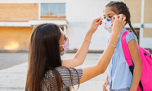 Mamá ayudando a una niña con mascarilla a prepararse para ir a la escuela - Children's Health